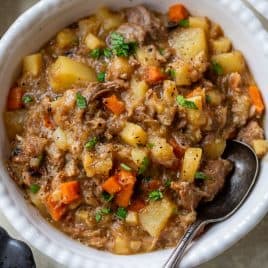 A bowl of pork stew with potatoes and carrots, garnished with fresh herbs and served with a metal spoon.