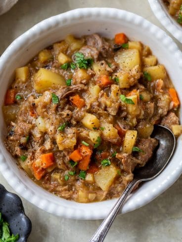 A bowl of pork stew with potatoes and carrots, garnished with fresh herbs and served with a metal spoon.