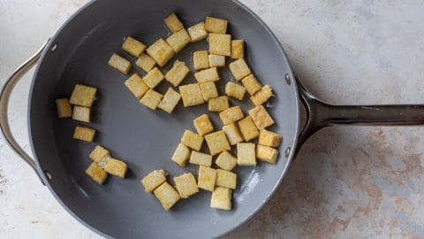 Cubes of golden-brown pan-fried tofu in a grey skillet.