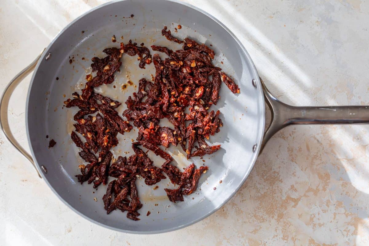 Sun-dried tomatoes and minced garlic cooking in a grey skillet.