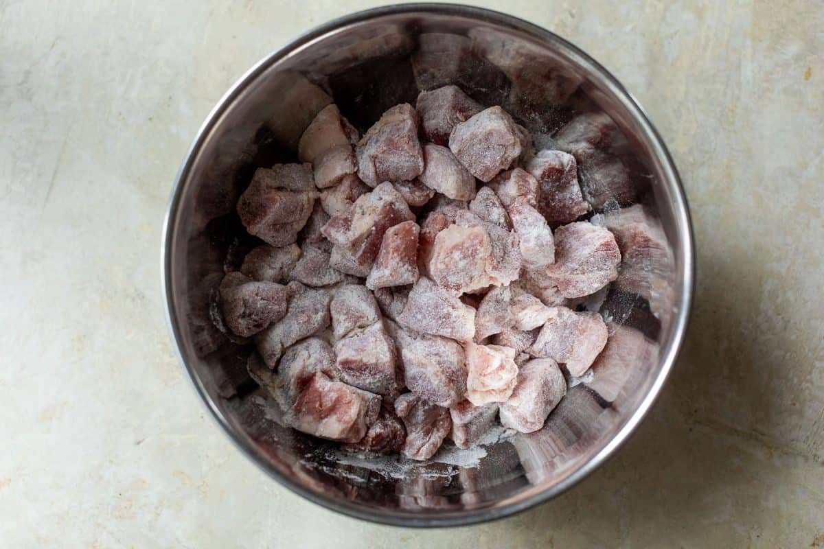 Metal mixing bowl filled with raw, flour-dusted pork cubes.