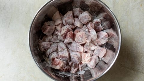 Metal mixing bowl filled with raw, flour-dusted pork cubes.
