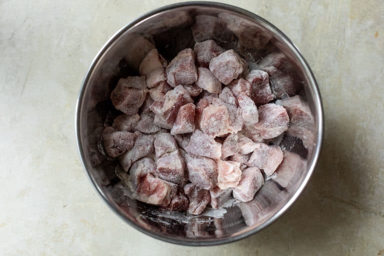 Metal mixing bowl filled with raw, flour-dusted pork cubes.