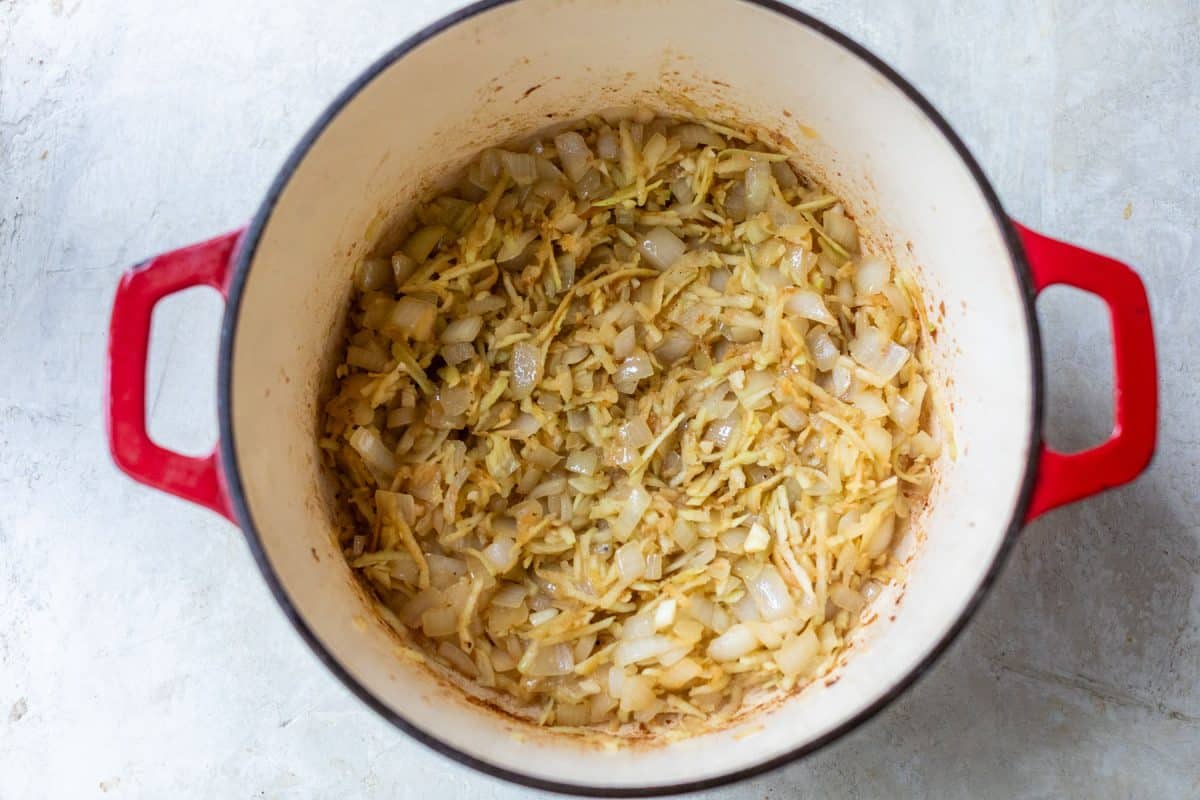 Chopped onions and shredded vegetables sautéing in a red enameled cast iron pot.