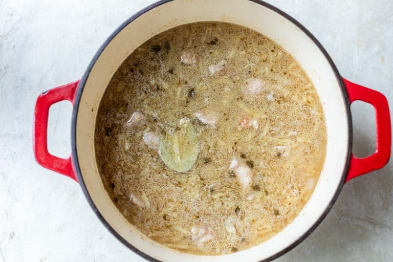 A red enameled cast iron pot filled with a light-colored savory stew containing meat and a bay leaf.