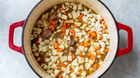 A red Dutch oven filled with diced potatoes, carrots, and chunks of pork in a savory broth.