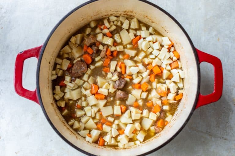 A red Dutch oven filled with diced potatoes, carrots, and chunks of pork in a savory broth.