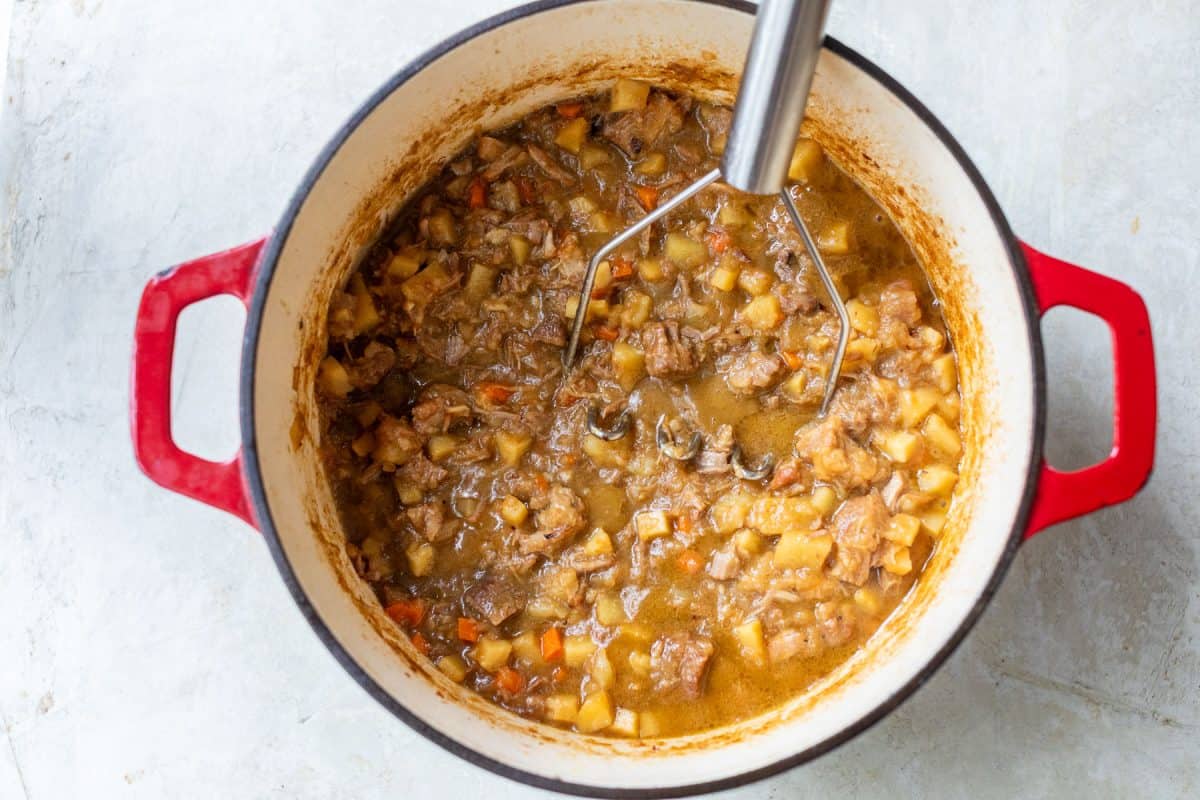 A potato masher in a red enameled Dutch oven filled with a chunky pork stew.