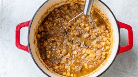 A potato masher in a red enameled Dutch oven filled with a chunky pork stew.