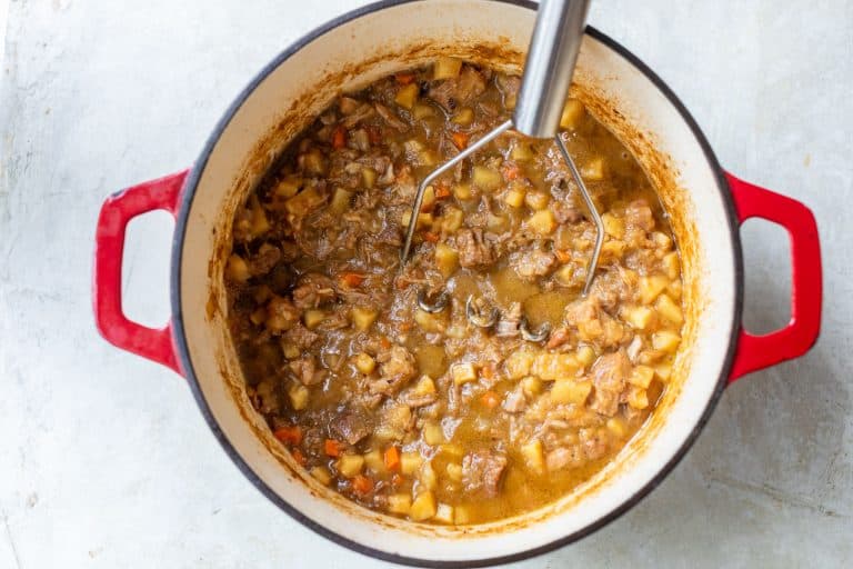 A potato masher in a red enameled Dutch oven filled with a chunky pork stew.