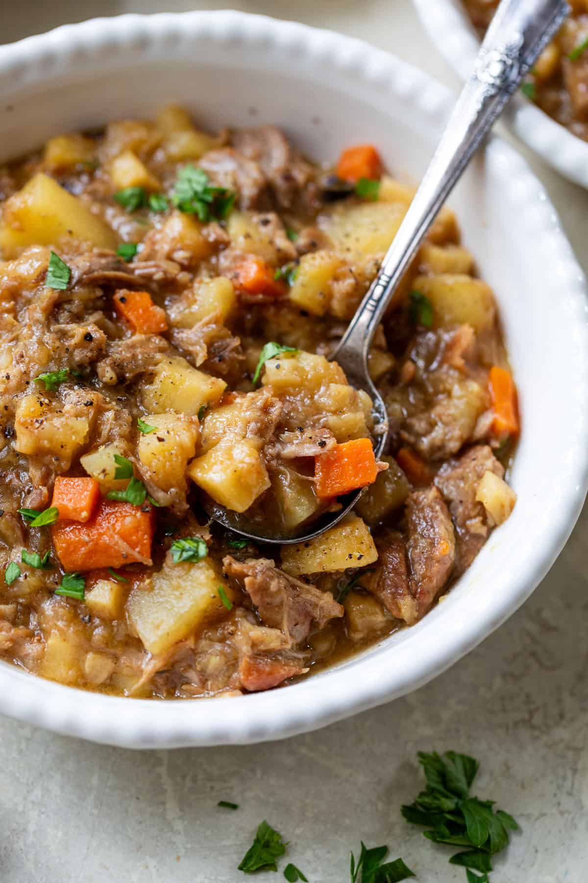 A spoon scoops pork stew with potatoes and carrots from a white bowl.
