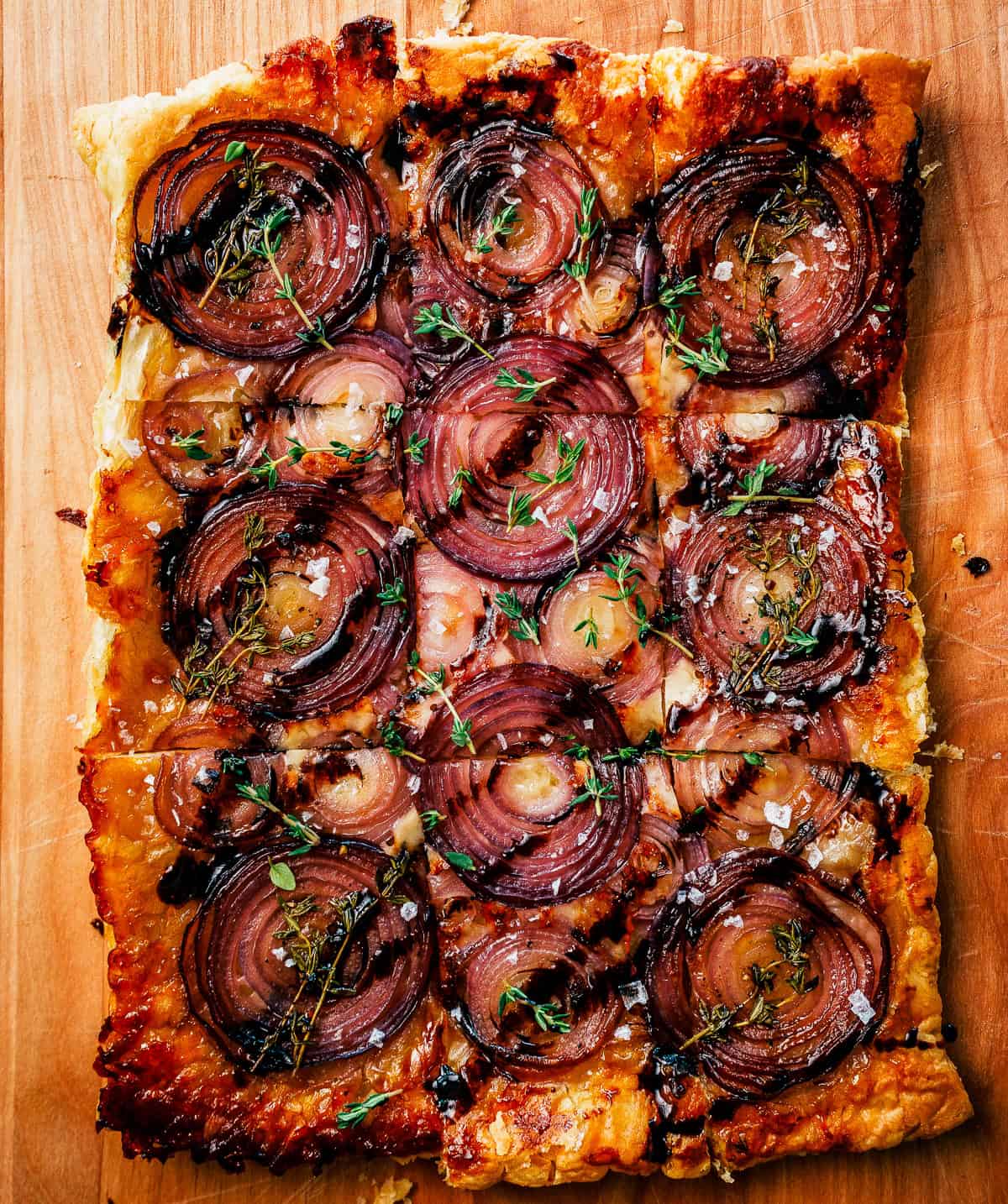 Rectangular onion tart sliced into squares and garnished with fresh thyme on a wooden board.